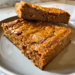 ['Close-up side view of healthy pumpkin blondies with chocolate chips.', 'Delicious gluten-free pumpkin blondies on a textured surface.', 'Juicy pumpkin blondies featured with white chocolate chips.', 'Tasty fall treat, pumpkin blondies with a golden-brown color.']