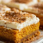 ['Close-up view of classic pumpkin bars topped with cream cheese frosting.', 'Sliced pumpkin bars with a smooth cream cheese layer, highlighted in natural light.', 'Side view of moist pumpkin bars, adorned with a dusting of powdered sugar.', 'A delicious serving of Kürbis bars with creamy frosting on a rustic surface.']