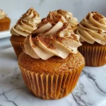 ['Close-up of vegan pumpkin cupcakes topped with cream cheese frosting and decorative Biscoff pumpkins.', 'Side view of moist vegan pumpkin cupcakes showcasing their fluffy texture and autumn-themed decorations.', 'A collection of vegan pumpkin cupcakes with a light brown frosting and pumpkin spice sprinkles.', 'Delicious vegan pumpkin cupcakes arranged neatly, featuring a thick layer of frosting and festive pumpkin toppers.']