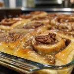 Close-up view of a cooked cinnamon roll casserole topped with icing and chopped pecans.